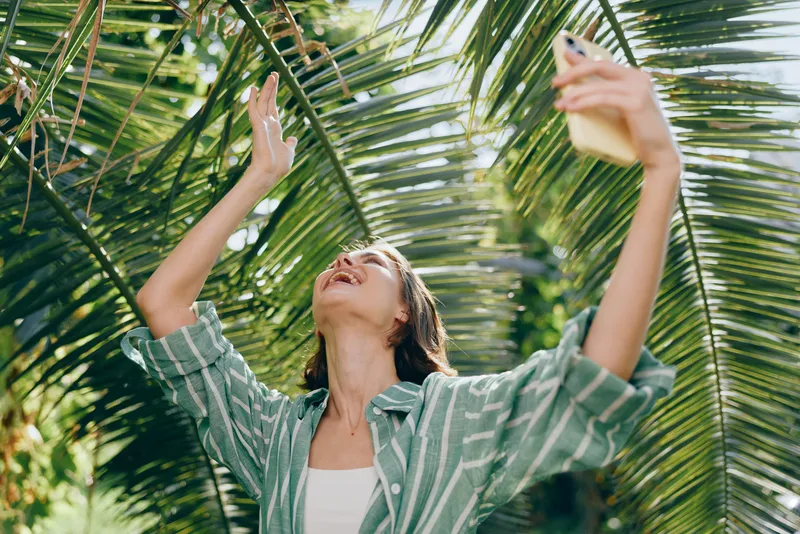 Woman in a striped shirt stands beneath bright palm fronds, raising her arms to selfie with a smartphone in a sunny outdoor setting.