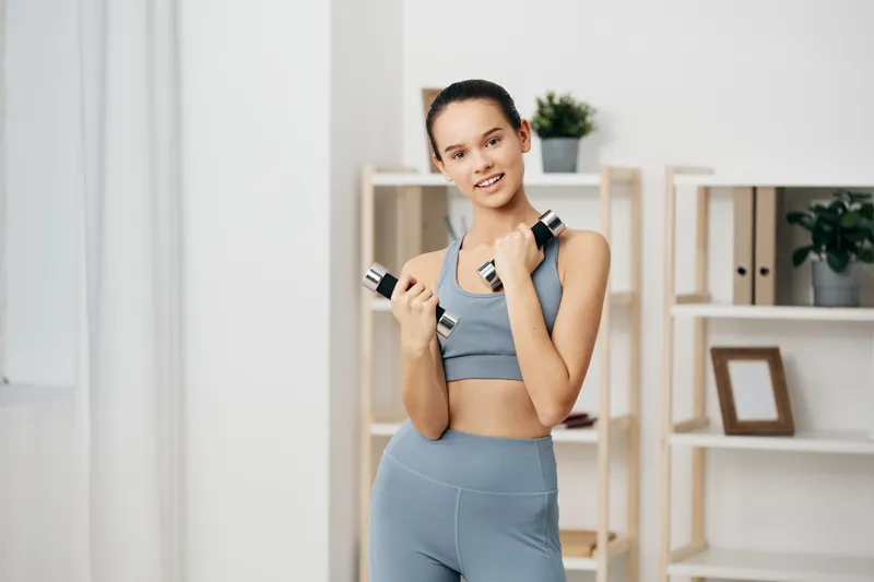 Fitness woman in sportswear holding dumbbells in a bright home studio, ready for workout and training. Clean shelves, minimal decor, focused expression, energy and health in a modern living space.