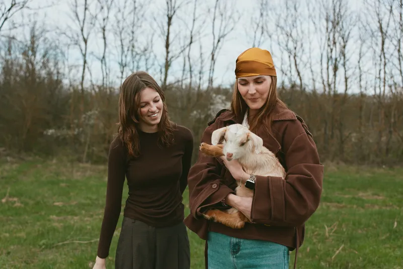 Goat, kid, lifestyle, outdoor, nature, woman, girl, friendship, field, meadow, sunlight, smiles, relaxed, clothing, jacket, cap, scarf, breeze, laughter, moment, candid, countryside, scenery, rustic,