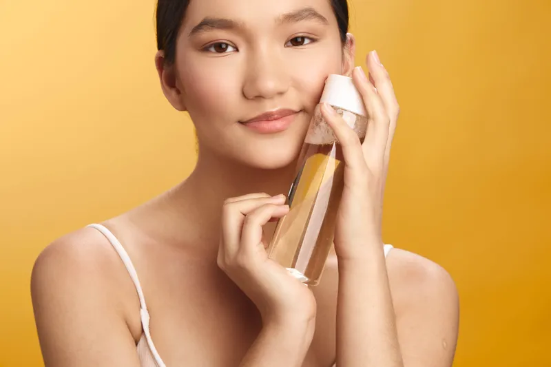 Asian woman holds a bottle against a warm yellow backdrop, smiling softly. The scene conveys elegance, freshness, and beauty in a clean, modern lifestyle setting.