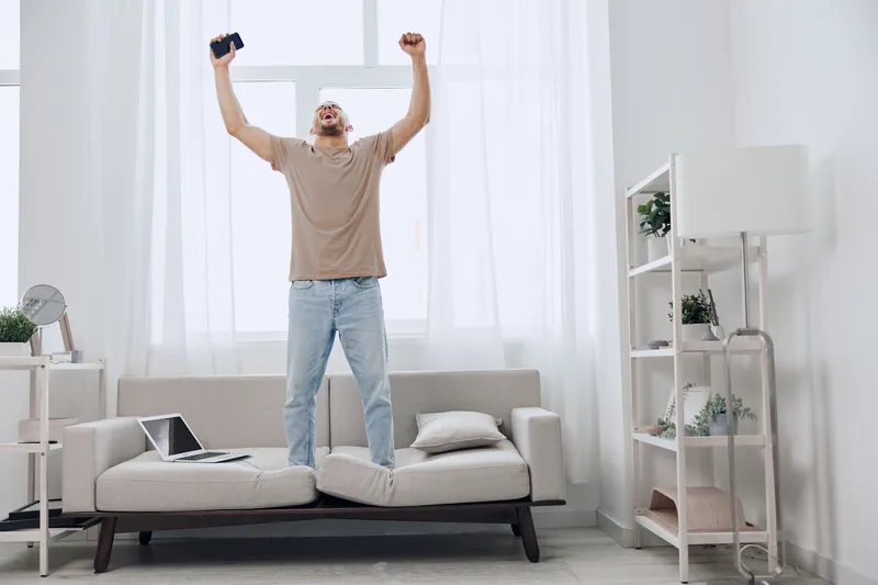 Energetic man celebrating success while standing on a sofa in a bright, modern living room. His excited expression showcases the joy of achievement and inspiration.