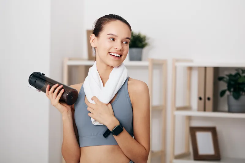 Woman fitness athlete at home, lifting a dumbbell with one hand, towel around her neck, smartwatch on wrist, focused expression, calm indoor backdrop.