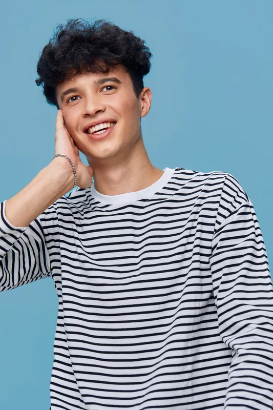 Smiling man in striped shirt posing against blue background, youthful expression, casual style, curly hair, joyful mood, studio portrait composition