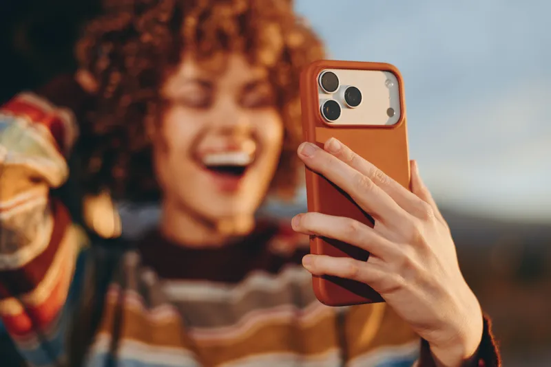 Woman with curly hair laughing, holding smartphone for a casual selfie outdoors, warm sunset light, shallow depth of field, lifestyle moment, friendly travel atmosphere, bright mood