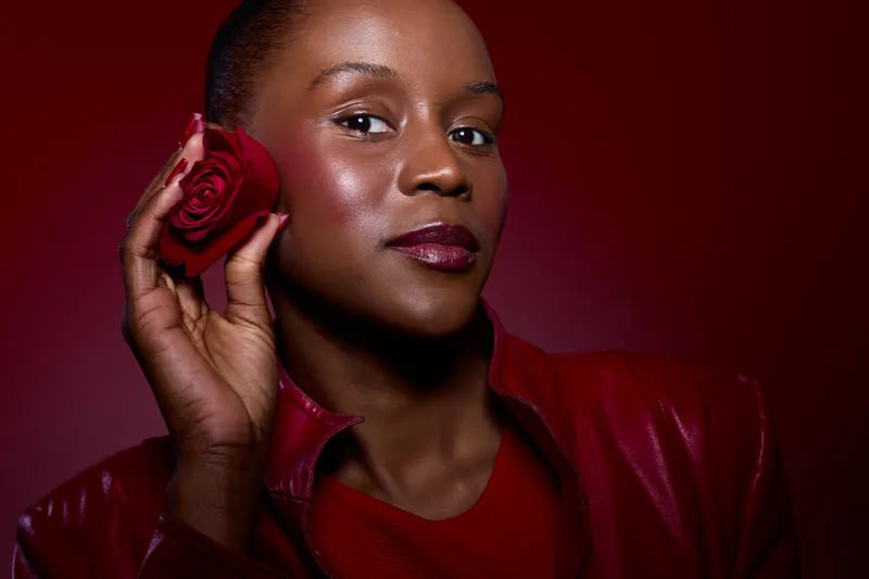 Confident dark skinned African woman holds red rose near face, wearing red leather jacket and lipstick. Studio portrait on dark red background. Concept of beauty, elegance, and self-expression.