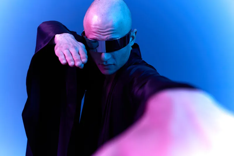 A striking man in a dark robe, wearing futuristic sunglasses and striking a dynamic pose against a vivid blue background, conveying a sense of mystery and intrigue.
