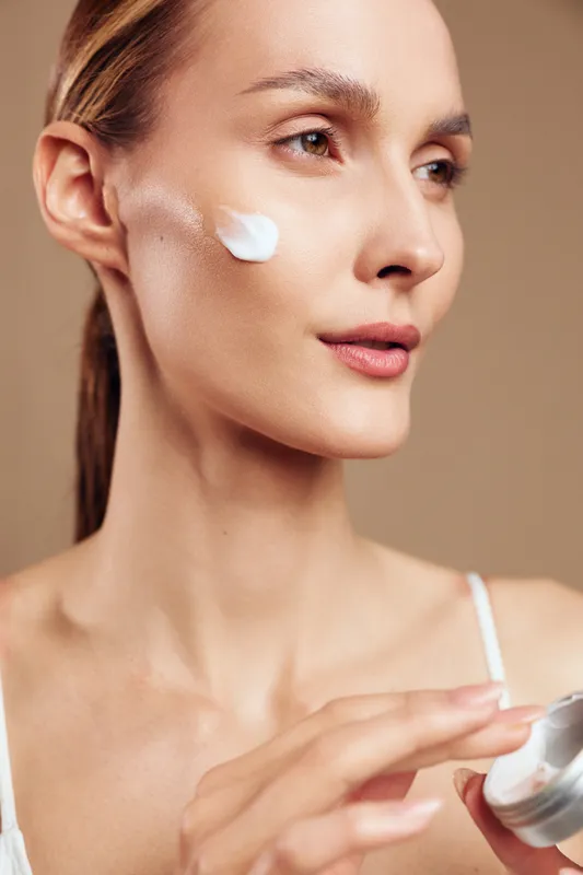 Young woman with clear skin applies moisturizing cream on her cheek while holding a jar of skincare product against a neutral background.