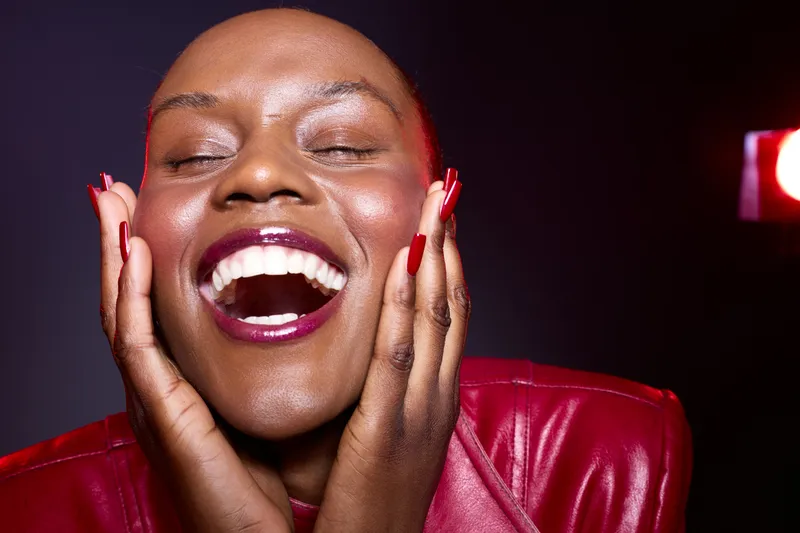 Joyful dark skinned African woman with short hair laughs with eyes closed, hands on face. Studio portrait with red jacket and bright lipstick. Concept of happiness and beauty.
