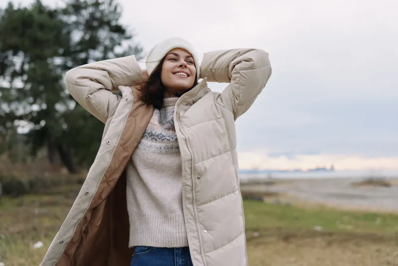 Woman outdoors wearing a puffy coat and knit sweater, smiling with hands behind the head, windy day by the coast, casual fashion and relaxed mood in nature and sea breeze.