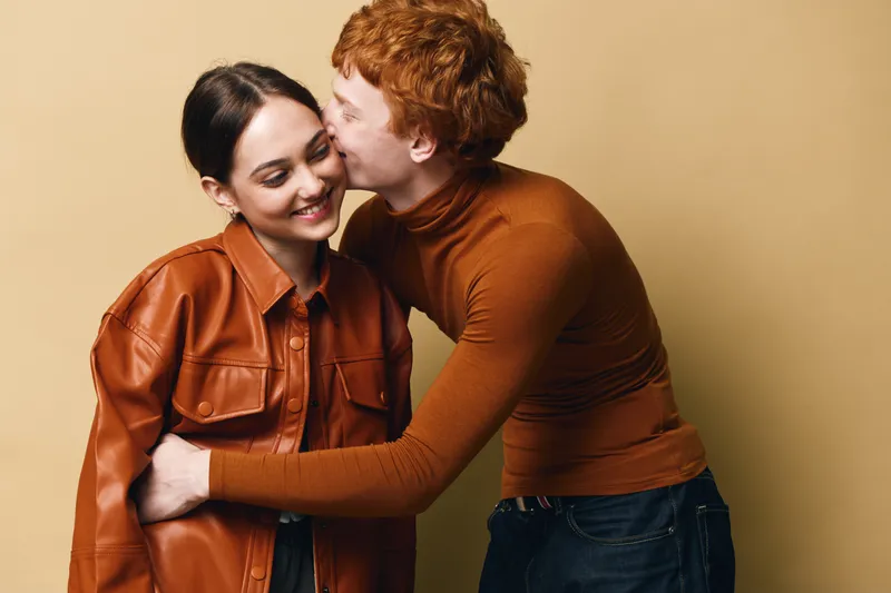 Couple embracing and smiling with affection in studio, young man kissing woman on cheek, warm clothing and casual style for love happiness and relationship.
