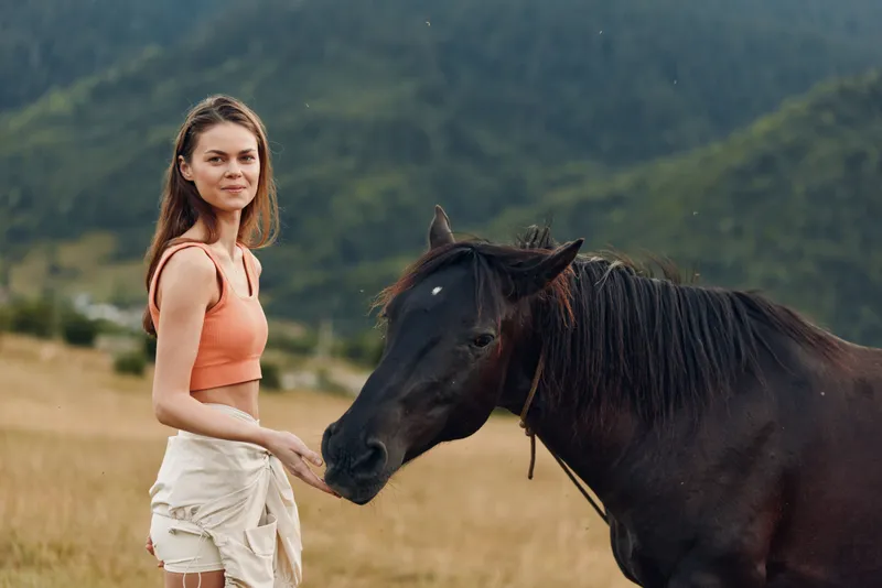 A young woman wearing an orange top and white shorts stands in a open field as a dark horse approaches. She looks calm and curious, sharing a gentle, peaceful moment with the animal.