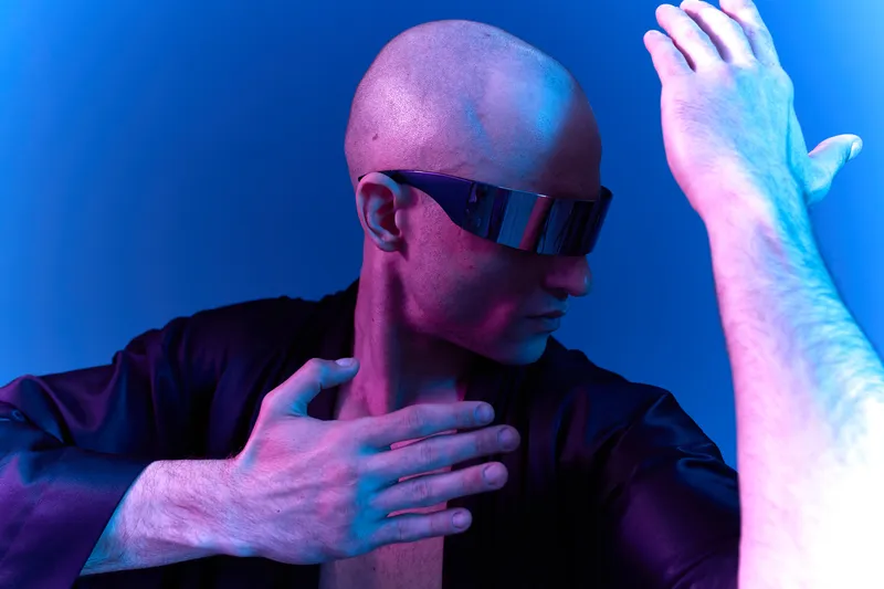 Stylish man with sunglasses posing dramatically against a vibrant blue and purple background. The image features modern aesthetics and a unique style expression.