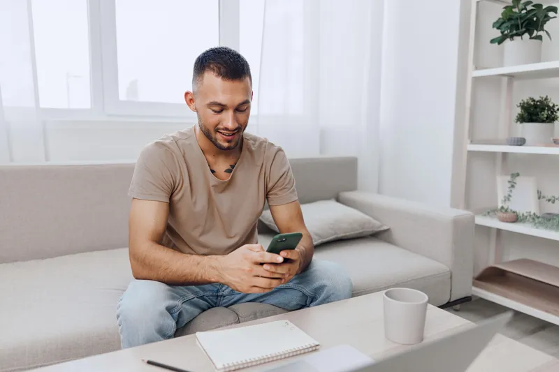 Smiling man enjoying leisure time at home while using a smartphone, dressed casually in comfortable clothing, surrounded by a cozy living space with natural light.