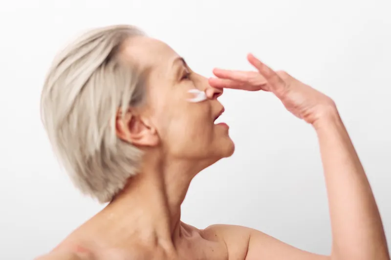 Mature woman with short blonde hair applies face cream on nose in bright studio. Skincare concept, healthy skin, anti aging routine, natural beauty, wellness and self care theme.