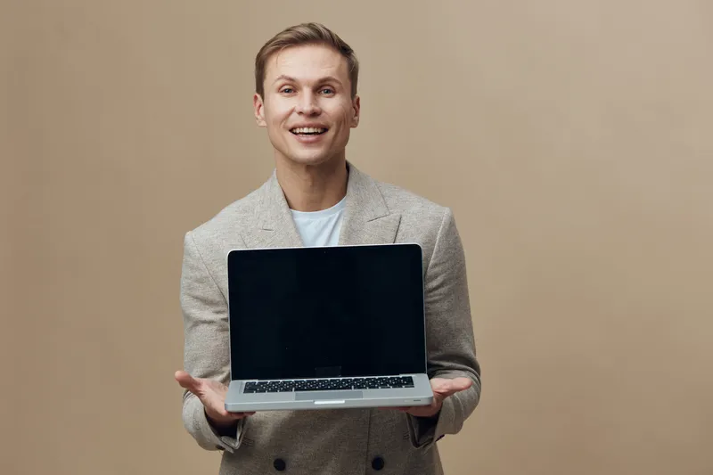 Smiling man with light skin, dressed in a beige blazer, holding a laptop with a black screen, standing against a plain beige background. Studio portrait of a professional businessperson, technology, s