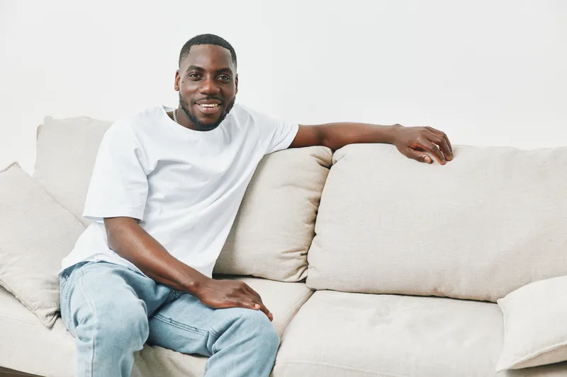 Young African American man relaxing on a light colored sofa, wearing a casual white t shirt and jeans, promoting a comfortable lifestyle and home atmosphere