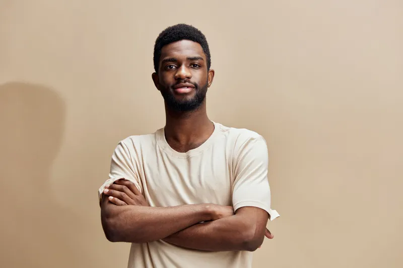 Confident young African man in beige t shirt with arms crossed against a warm background, expressing self assurance and positivity