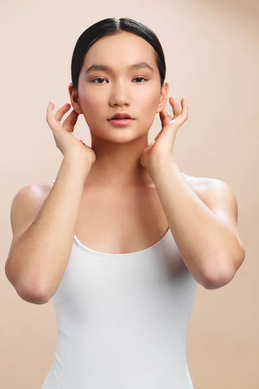 asian woman wearing a simple white tank offers a calm, intimate studio portrait with soft beige background, showcasing natural beauty, balance, and subtle confidence.