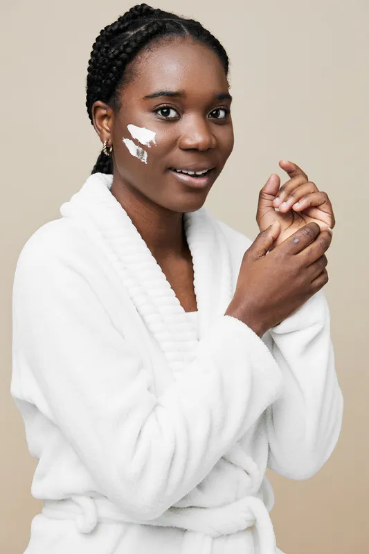 Young Black woman applying skincare product with a gentle smile in a white bathrobe against a soft beige background, promoting beauty and self care