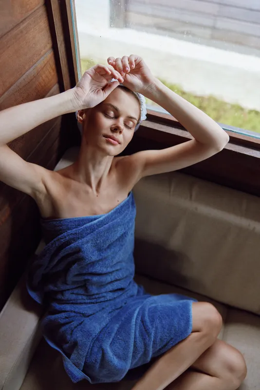 Relaxed woman wrapped in blue towel resting by wooden window with closed eyes, enjoying peaceful moment and natural light, serene atmosphere, calm skin care routine.