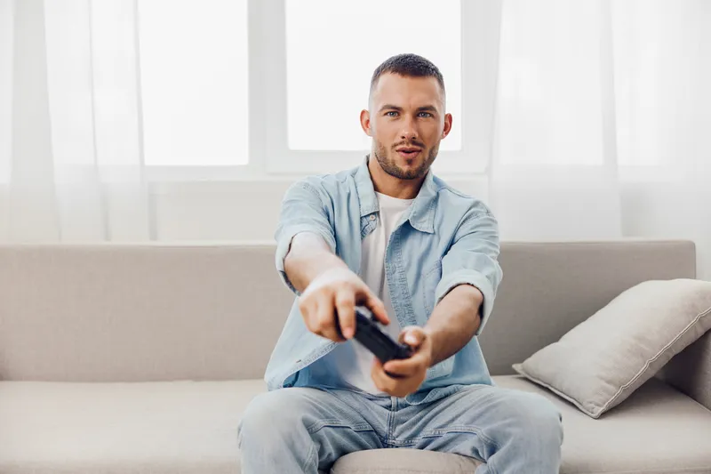 Engaged man enjoying leisure time at home, holding a remote control, relaxing on a stylish couch with bright natural light streaming through the window.