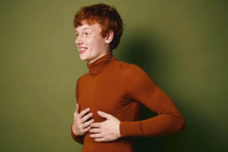 Young man with red hair wearing brown turtleneck sweater posing smiling and touching chest against green background in studio. Casual portrait with relaxed mood and confident expression.