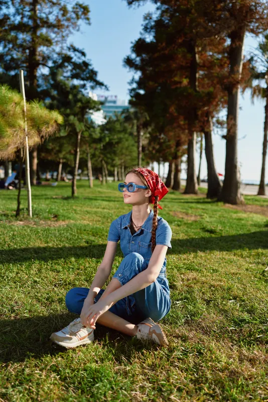 Lifestyle woman in modern boho-western denim street style sitting on grass outdoors with sunglasses, red bandana, and relaxed posture under sunny sky.