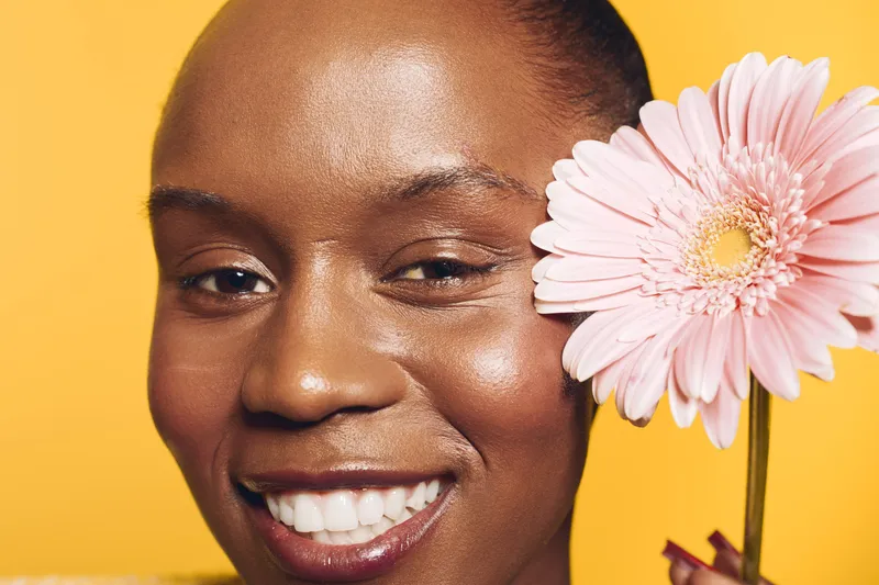 Close-up of a happy African woman with dark skin holding a pink gerbera flower near her face. Bright yellow background, natural beauty, skincare, wellness, and positive mood concept.