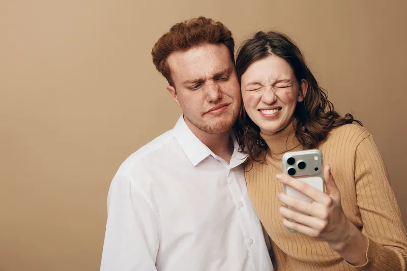 Young couple making funny faces while taking a selfie with smartphone on beige background. Casual clothing, playful mood, and close relationship concept.