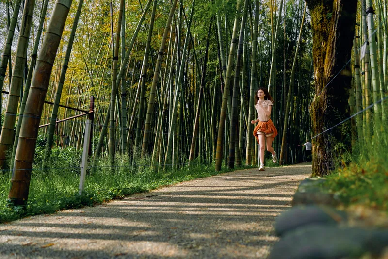 Woman running on a shaded bamboo forest path for nature and exercise, young female jogger in summer outfit, motion and fitness on a gravel trail surrounded by tall green bamboo.
