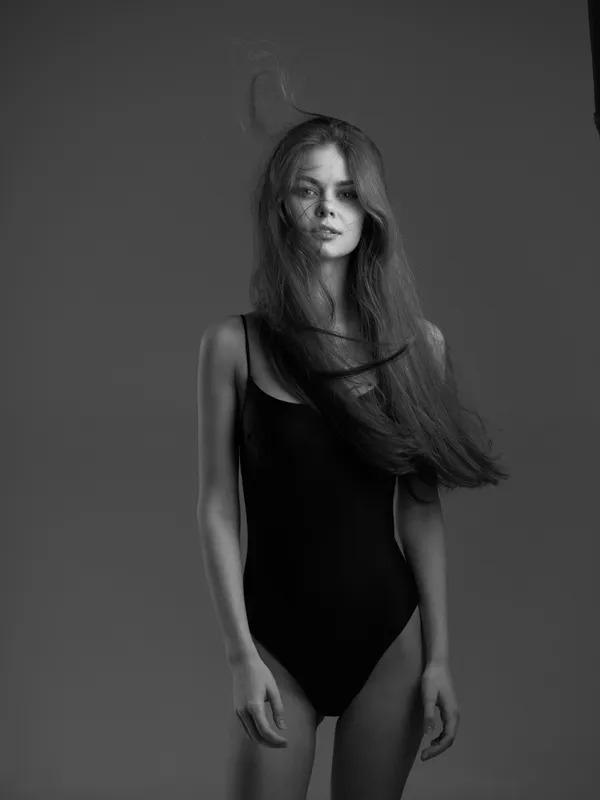 Black and white portrait of a young woman with long hair wearing a black bodysuit, standing against a plain gray background
