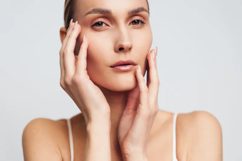 Close-up portrait of a young woman with clear skin touching her face gently. Soft lighting highlights natural beauty and skincare concept on a plain white background.