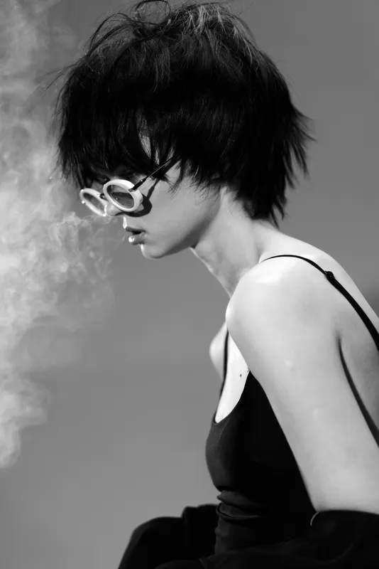 Black and white portrait of a woman with short hair wearing round glasses and a dark tank top in a smoky atmosphere