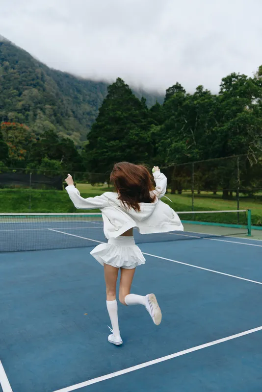 Young female athlete enjoying a joyful moment on a tennis court, wearing a sporty white outfit with a backdrop of lush green mountains and moody skies