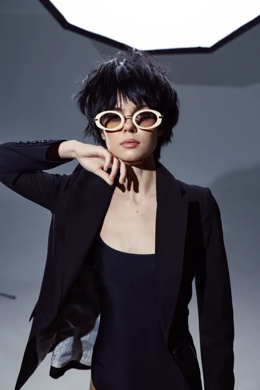 Stylish woman in black blazer and white sunglasses posing with confident expression in studio lighting.