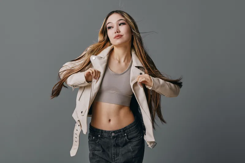 Young woman with long hair posing in beige jacket and crop top under studio light on gray background. Fashion, confident female model wearing casual style with trendy outfit and natural look.