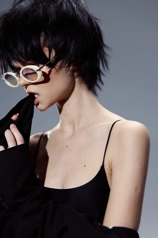 Stylish woman wearing round glasses biting black fabric with intense expression and short hair.