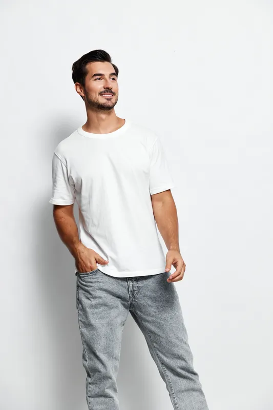 Full body smiling happy cheerful young man wearing a white t-shirt and gray pants, looking confidently at the camera against a plain light gray background.