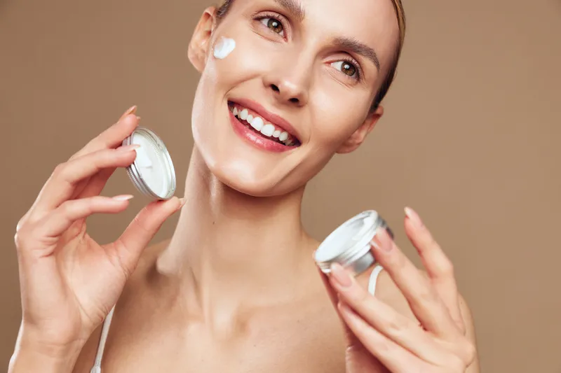 Smiling woman applies cream on face holds jar of skincare product shows healthy glowing skin with natural makeup isolated on beige background bright positive beauty concept.