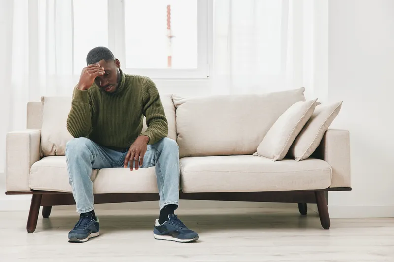 A distressed African American man sitting on a sofa, holding his head in his hand, wearing a green sweater and jeans, expressing feelings of sadness and anxiety