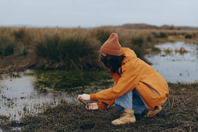 Outdoor lifestyle portrait of a woman kneeling on a riverbank, wearing orange coat, straw hat, and boots, hands touching soil, peaceful soft rural nature landscape with water and reeds.