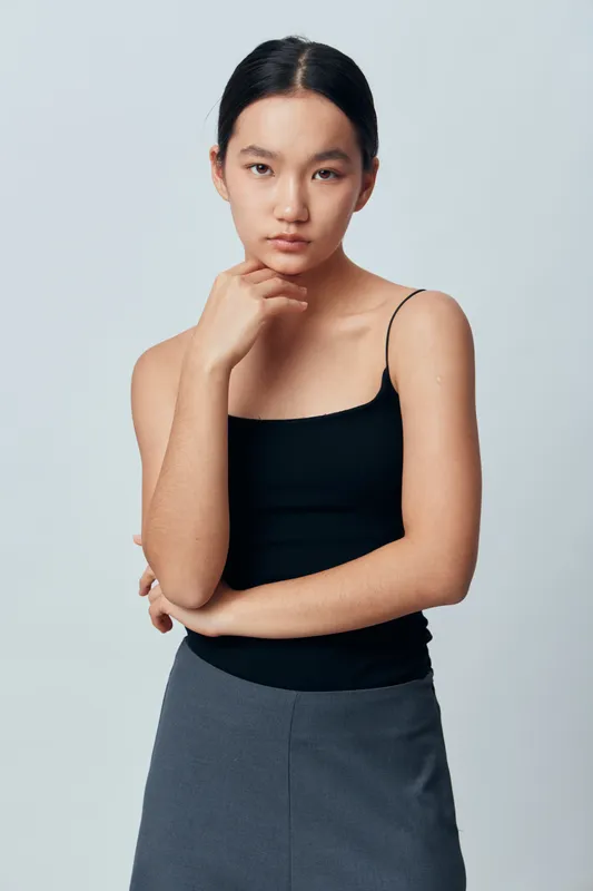 Young woman with black hair and natural makeup wearing black tank top and gray skirt posing thoughtfully on light gray background in studio. Portrait with serious expression.