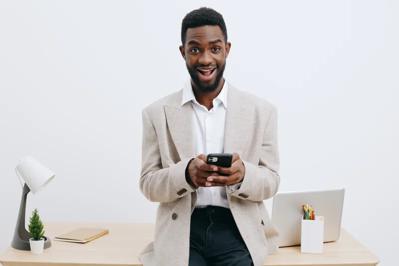 Smiling young man with dark skin in a beige blazer and white shirt holding a smartphone, standing in a modern office with minimalistic decor and a laptop on the desk. People lifestyle concept, technol