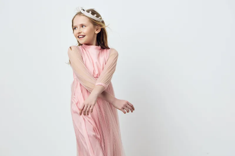 Cheerful girl in a pink dress with a crown, showcasing joy and playfulness against a light background for children s fashion and portraits