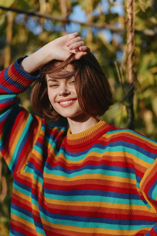 Young woman in a striped sweater smiles in a sunlit outdoor nature portrait, short brown hair, casual outfit, hand shading eyes, cheerful lifestyle fashion portrait.