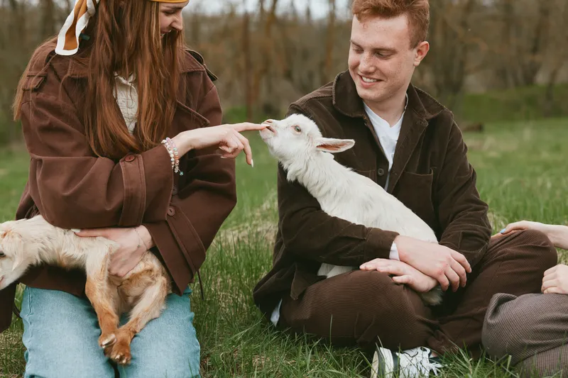 Cheerful outdoor scene with two friends, a man and a woman, gently interacting with a playful goat kid in a sunlit meadow, casual clothes, relaxed atmosphere, natural colors, candid moment of