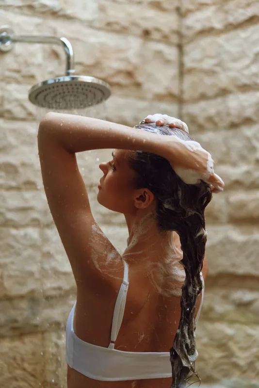 Woman washing long hair with foam in indoor shower, natural stone background, soft light, relaxing self-care moment, wellness routine, personal hygiene.