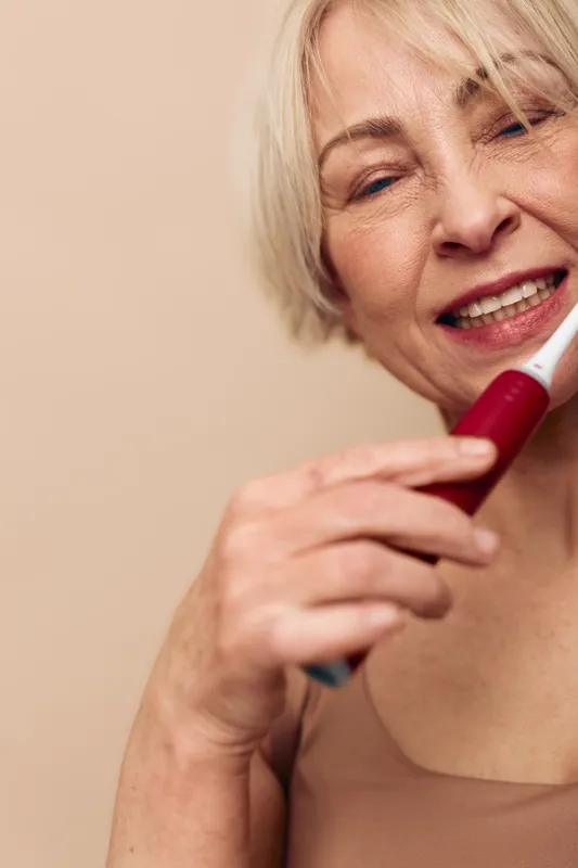 Close-up of smiling mature woman holding electric toothbrush, demonstrating dental care routine at home with natural light. Concept of oral hygiene, healthy teeth, and daily self-care.