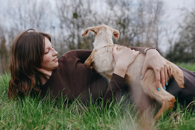 Woman lying on grass, cuddling a young dog in a tender moment. Outdoor setting conveys relaxation, affection, and simple lifestyle. Cozy, intimate scene with soft light and natural textures.