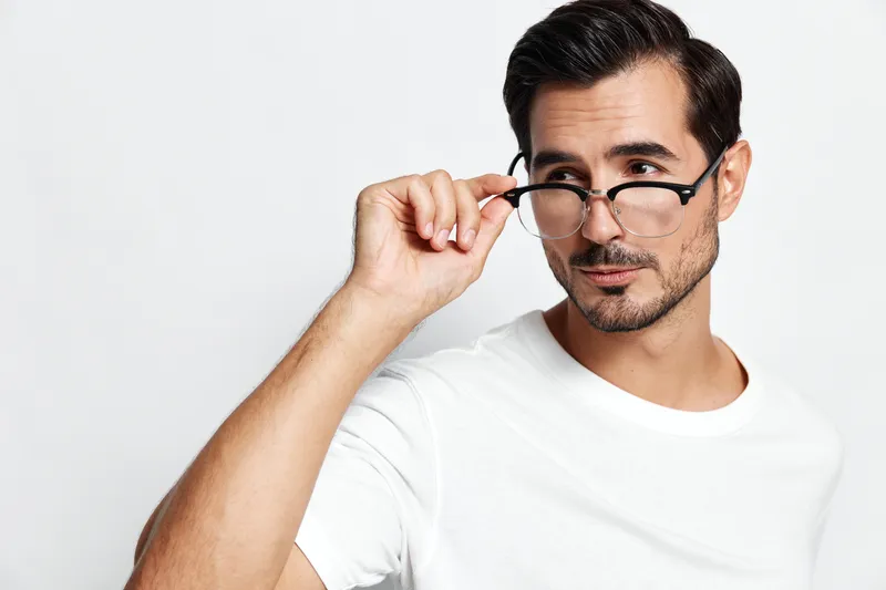Stylish young man wearing glasses and a white t-shirt, confidently looking at the camera while adjusting his eyewear, isolated on clean solid background.